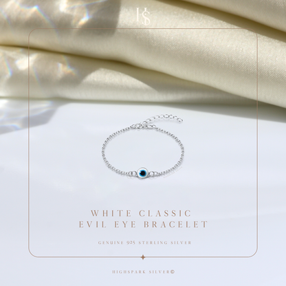 A sterling silver evil eye bracelet with a white and blue enamel charm, styled on a white surface with draped cream satin fabric.