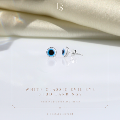 A pair of round evil eye stud earrings in sterling silver, styled on a reflective white surface with draped cream fabric in the background.