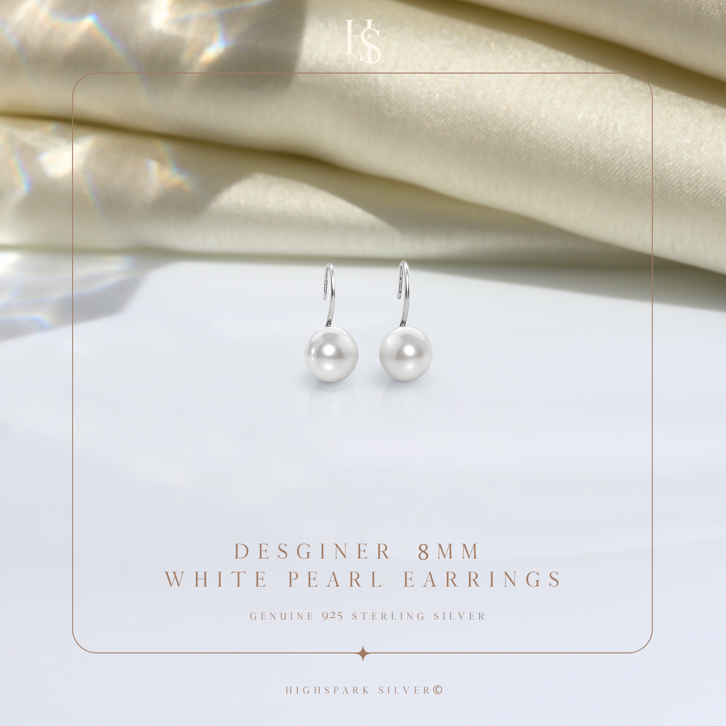 A pair of sterling silver hook earrings with round white pearls, styled on a white surface with draped cream fabric in the background.