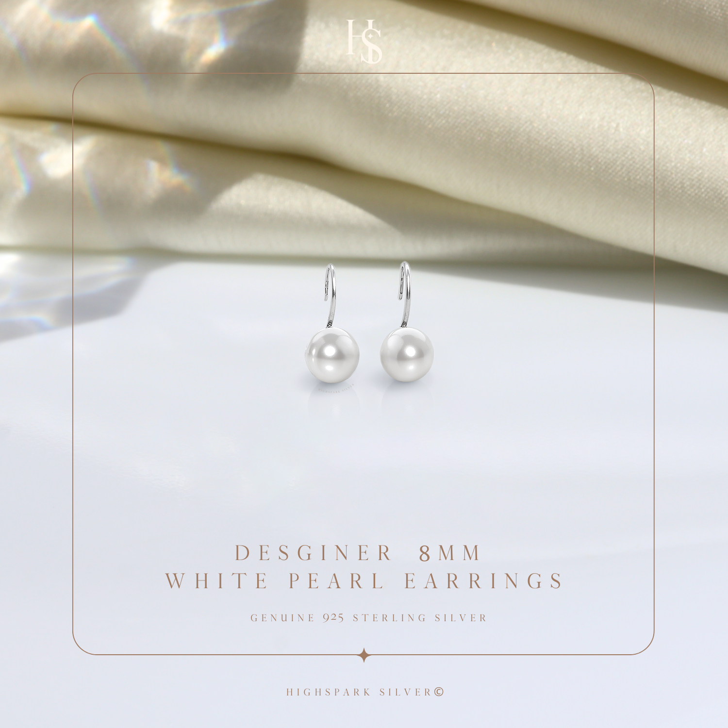 A pair of sterling silver hook earrings with round white pearls, styled on a white surface with draped cream fabric in the background.