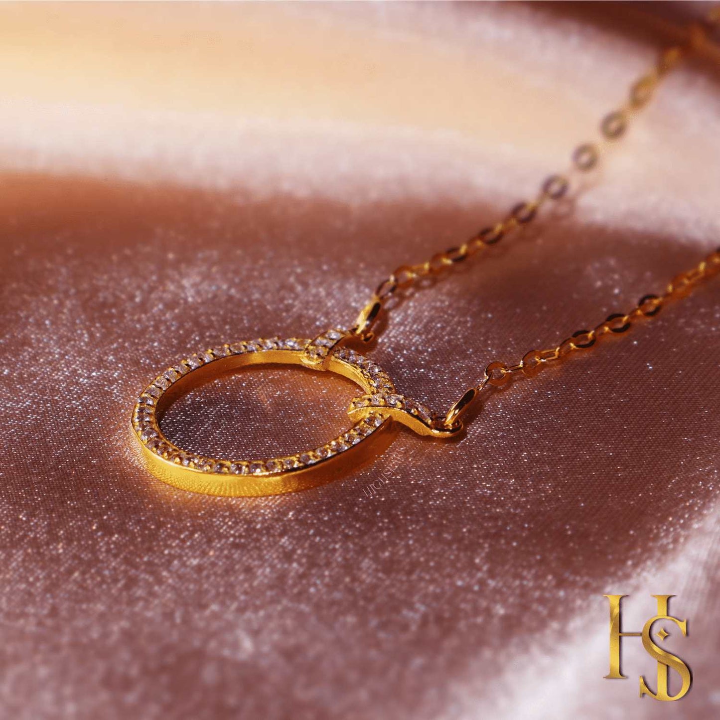 A gold-plated pendant necklace with a circular motif studded with American diamonds, styled on a shimmering pink satin surface.