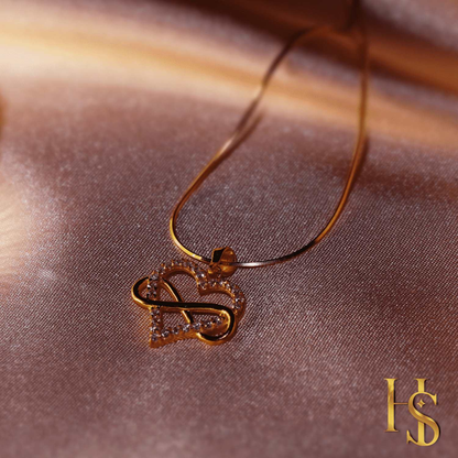 A gold-plated pendant necklace with an infinity symbol through a heart studded with American diamonds, styled on a dusty pink satin fabric.