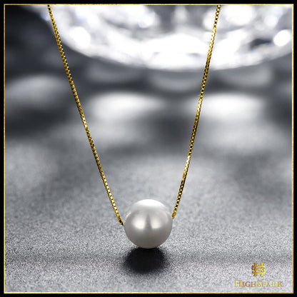 A single white pearl pendant on a gold-plated box chain necklace, styled on a textured grey surface with a soft, reflective background.