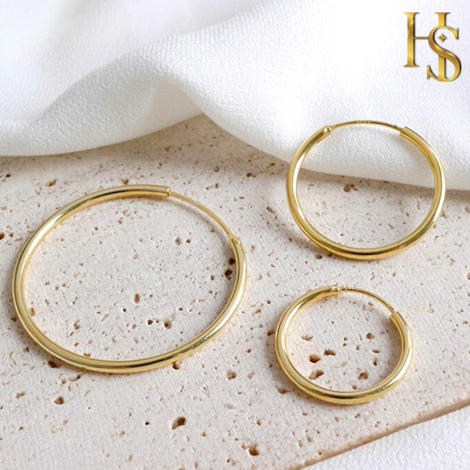 Three gold-plated sterling silver hoop earrings in different sizes are styled on a textured beige surface with white fabric.