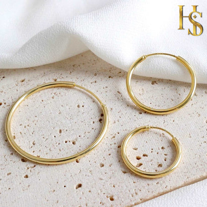 Three gold-plated sterling silver hoop earrings in different sizes are styled on a textured beige surface with white fabric.