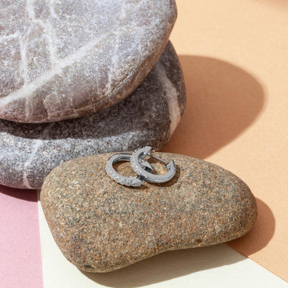 A pair of sterling silver hoop huggie earrings with pave-set American diamonds resting on smooth river stones.