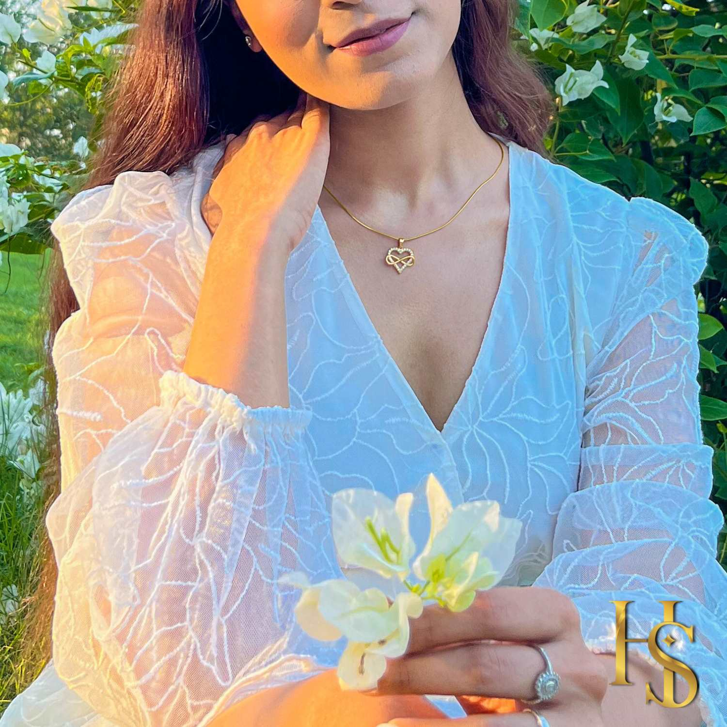 A gold-plated infinity heart pendant necklace with American diamonds, worn on the neck by a model in a white dress outdoors.