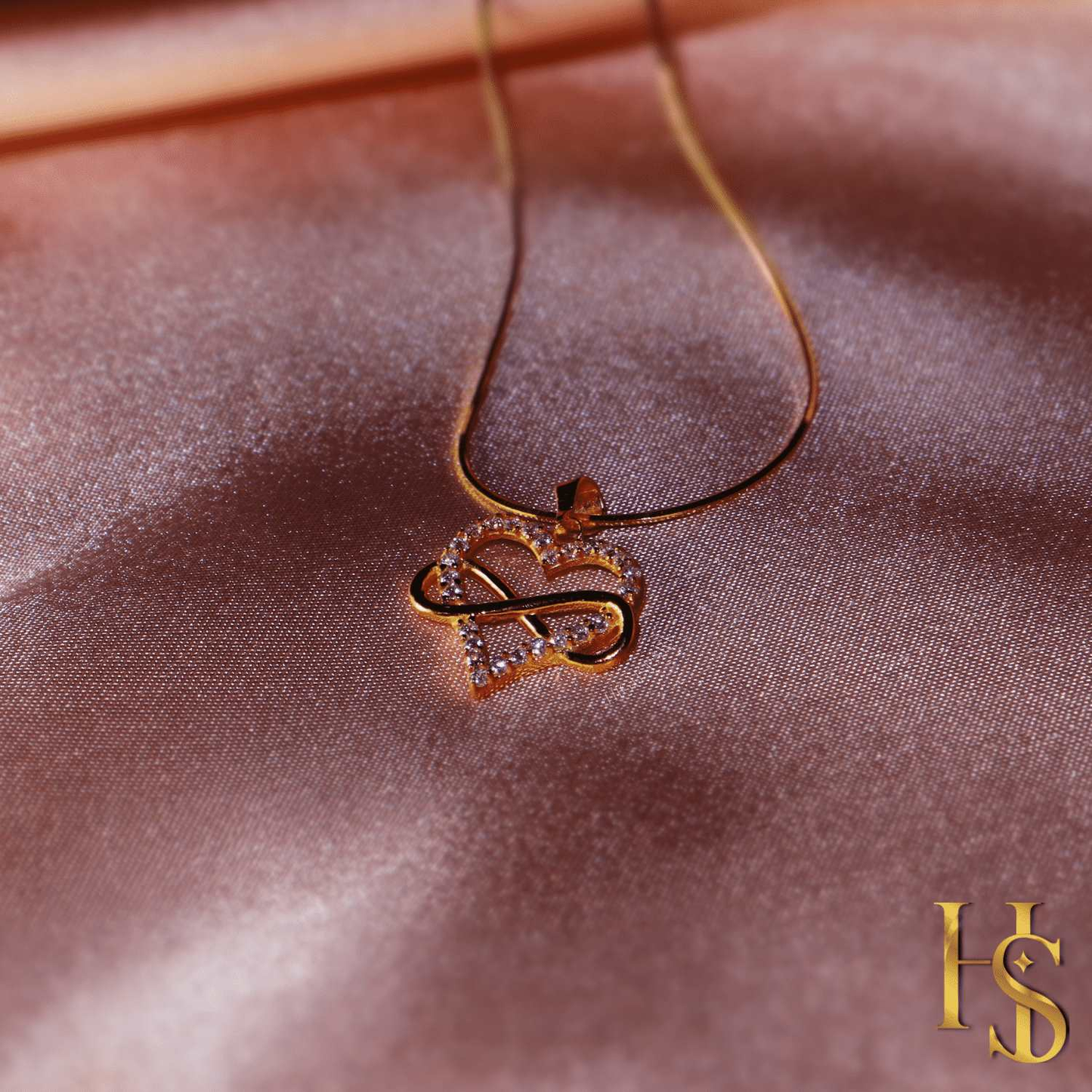 A gold-plated pendant necklace with a heart and infinity motif studded with American diamonds, styled on a shimmering pink satin surface.