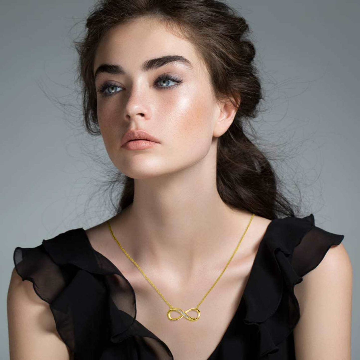 A gold-plated infinity pendant necklace, with one loop pavé-set with American diamonds, worn on the neck of a model in a black top.