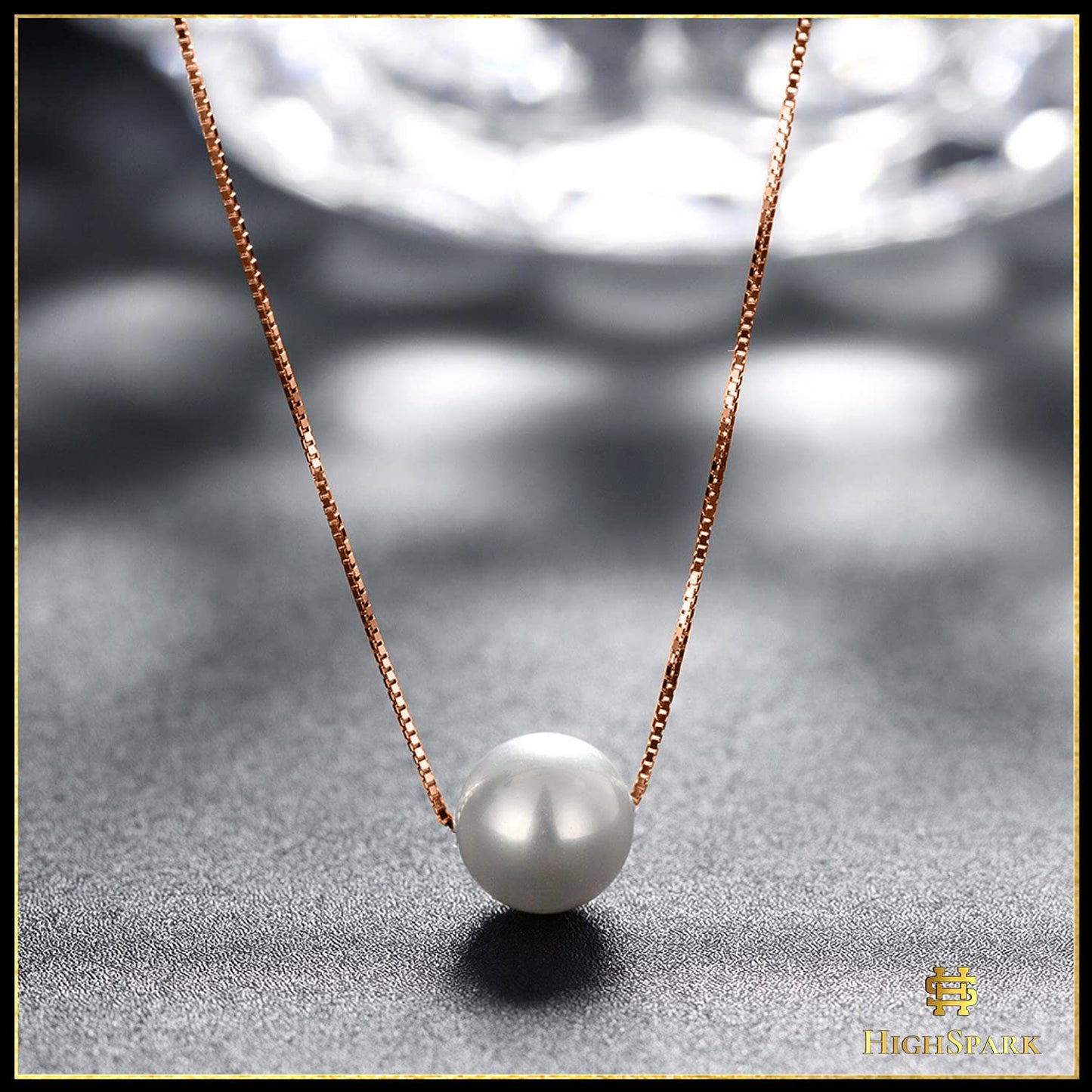 A single white pearl pendant on a rose gold-plated box chain, styled on a textured grey surface with a faceted glass background.