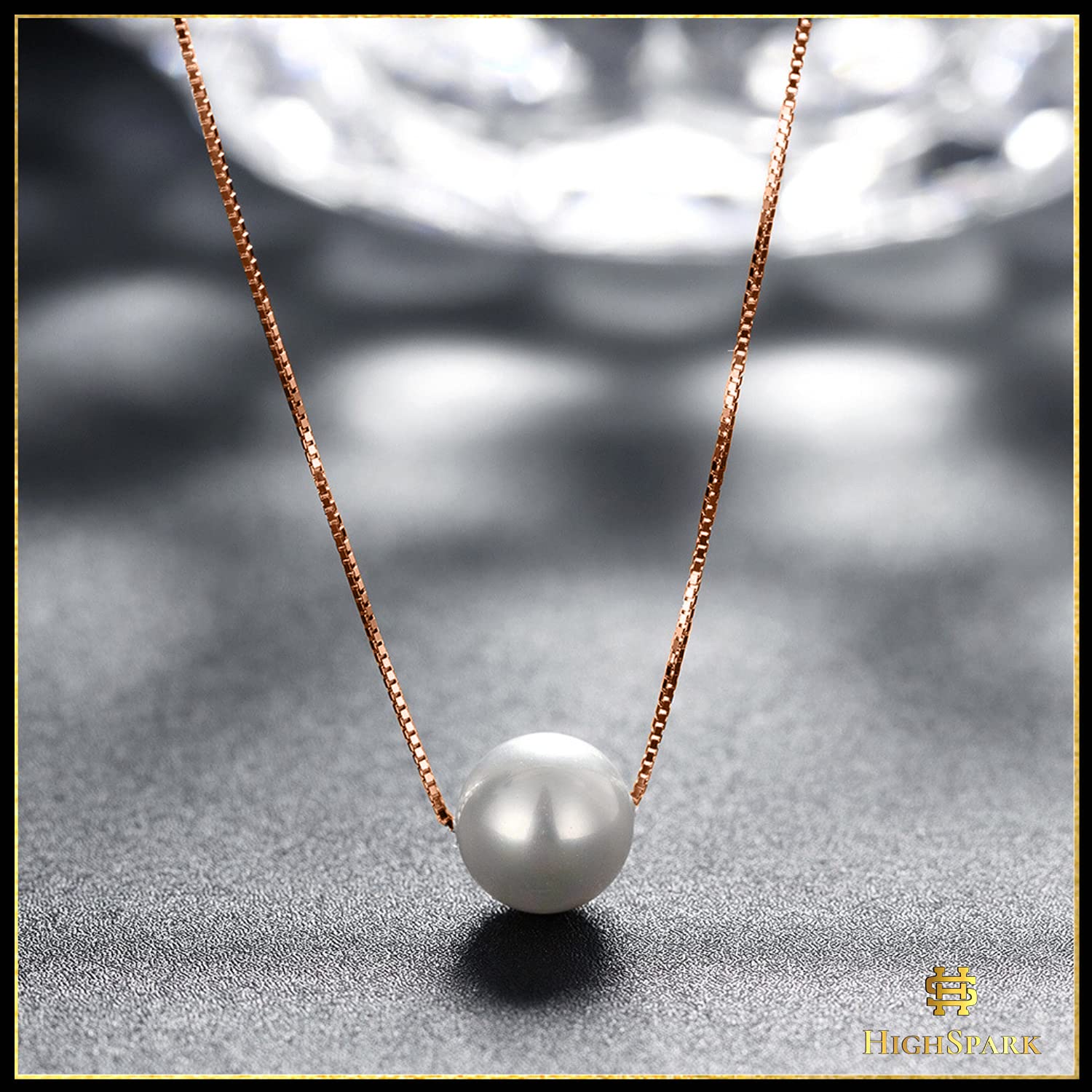 A single white pearl pendant on a rose gold-plated box chain, styled on a textured grey surface with a faceted glass background.